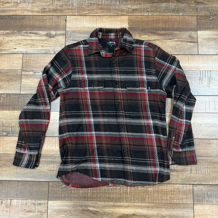 The Stussy Flannel Black/Red is a preowned long-sleeved shirt showcasing a classic blend of black, red, and white plaid, displayed flat on a wooden floor.