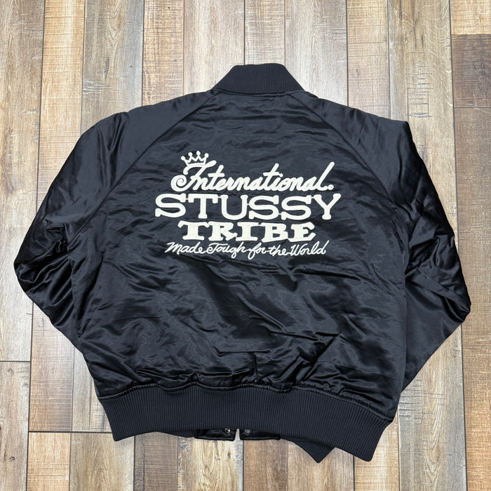 The Stussy Souvenir Jacket FW24 by Holy Ground Sneaker Shop is a black satin bomber with ribbed cuffs, collar, and hem. It features a snap button front and Stussy embroidered in white on the left chest. The jacket is laid flat on a wooden floor.