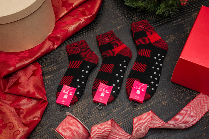Tall Order The Cabin Slipper Socks in black and red plaid with non-slip grips, flat lay surrounded by festive red wrapping materials.