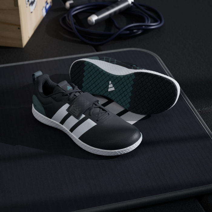 Adidas THE TOTAL 2 training shoes in black with white stripes, green heel accents, and a textured rubber outsole, angled view on gym mat.