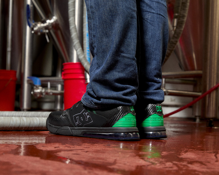 DC WorkCrew Versatile Work Comp Toe work shoes in black with green heel accents and breathable mesh material, worn in an industrial setting.