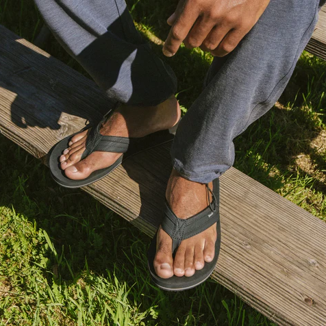 Sanuk Ziggy sandals in black with fabric straps and cushioned footbed, worn outdoors on wooden steps.